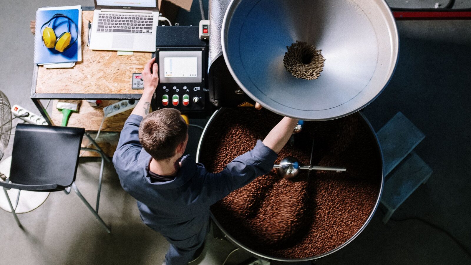 A roastery floor from above: drum roaster, cooling tray and a laptop logging the roast.